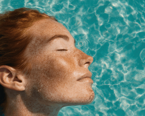 Primo piano del viso di una donna lentigginosa che galleggia in acqua con gli occhi chiusi, immagine evocativa dei segni lasciati dal sole sulla pelle e dell’importanza di trattare le discromie cutanee.