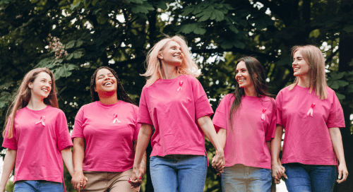 Cinque donne sorridenti camminano mano nella mano in un parco, indossando magliette rosa con il nastro simbolo della prevenzione del tumore al seno, durante un evento di sensibilizzazione per l’Ottobre Rosa.