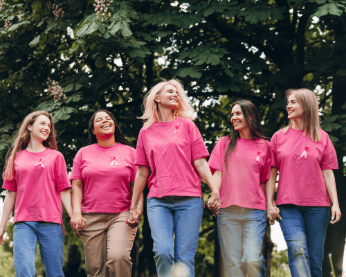 Cinque donne sorridenti camminano mano nella mano in un parco, indossando magliette rosa con il nastro simbolo della prevenzione del tumore al seno, durante un evento di sensibilizzazione per l’Ottobre Rosa.