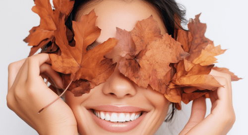 Donna sorridente con foglie autunnali sugli occhi, simbolo di benessere e cura della pelle nella stagione autunnale, rappresenta il concetto di dermocosmesi autunnale e rinnovamento cutaneo.