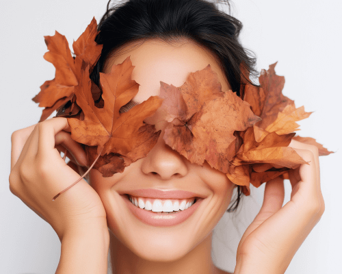 Donna sorridente con foglie autunnali sugli occhi, simbolo di benessere e cura della pelle nella stagione autunnale, rappresenta il concetto di dermocosmesi autunnale e rinnovamento cutaneo.