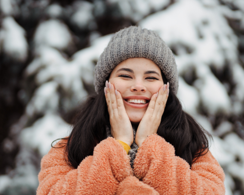 Donna sorridente con cappello di lana e giacca calda che si tocca il viso all’aperto, simbolo della cura della pelle contro il freddo.