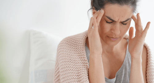 Donna seduta sul letto con le mani alle tempie e gli occhi chiusi, che mostra segni di dolore alla testa, simbolo della prevenzione e gestione di emicranie e mal di testa stagionali.
