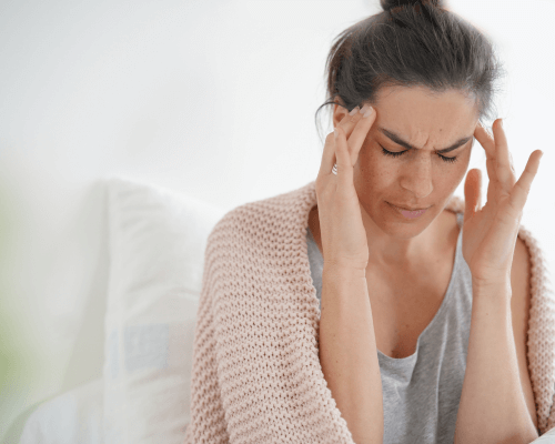 Donna seduta sul letto con le mani alle tempie e gli occhi chiusi, che mostra segni di dolore alla testa, simbolo della prevenzione e gestione di emicranie e mal di testa stagionali.