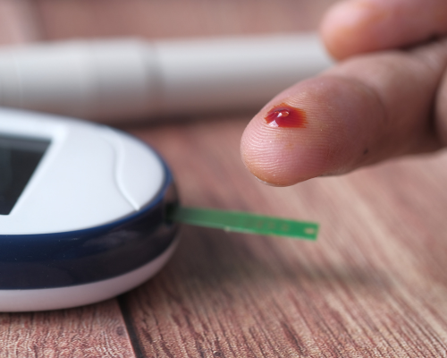 Primo piano di un dito con una goccia di sangue accanto a un glucometro pronto per la misurazione della glicemia.