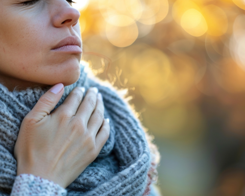 Donna avvolta in una sciarpa di lana che si tocca la gola, con espressione di fastidio, in un ambiente invernale.